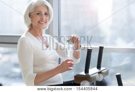 Fitness break. Joyful fit attractive woman relaxing and drinking water in the gym after working out.