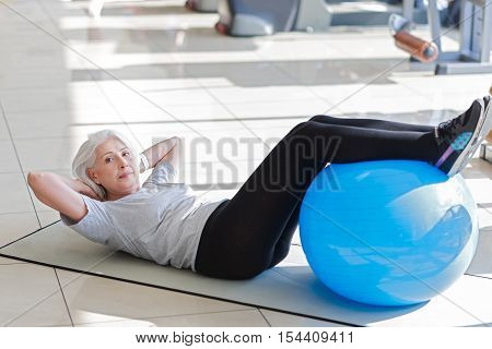 I will do it. Ambitious pretty senior woman laying on a floor and putting legs on a fit ball while doing sit ups.