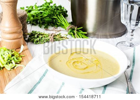 Classic healthy vegetable potato and leek soup based on vegetable broth served in white bowl with olive oil. White wood background. Steel pot with soup, parsley, peppermill and glass of water on back
