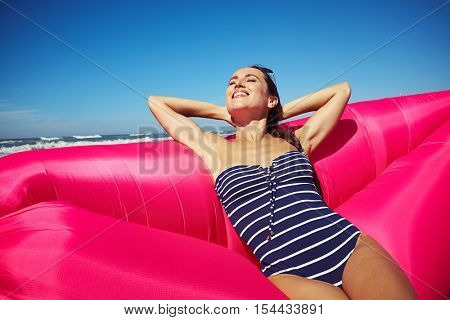 lady closing her eyes and keeping her hands beyond the head on the sunny beach, sitting on the deep rosy air rubber boat and having her moment while the sun is shining. 