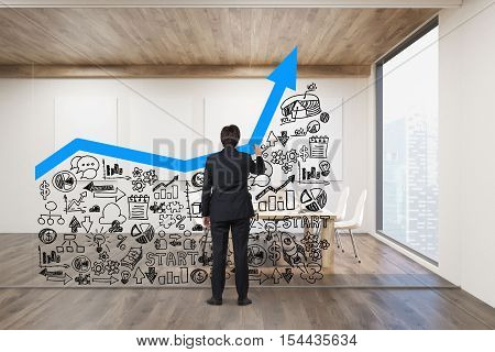Rear view of businessman drawing large blue graph and small business icons on glassboard in office. Concept of the right business strategy. Mock up. 3d rendering.