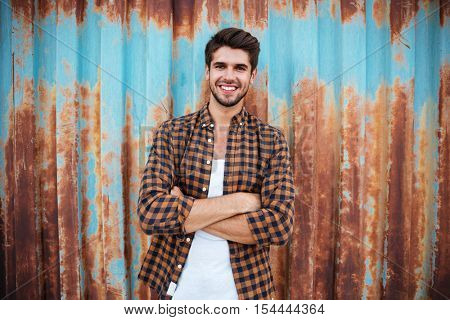 Happy young man in plaid shirt standing with arms crossed over blue metal background with rust