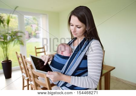 Beautiful young mother with her newborn baby son in sling at home, holding tablet, writing something