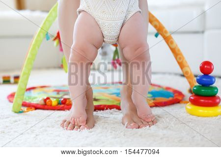 Bare pink feet of a little girl of 8 months standing on my mother's bare feet with a pedicure pink color,the girl dressed in panties white with light brown pattern,mom's legs standing on white soft rug in the nursery on the background of toys