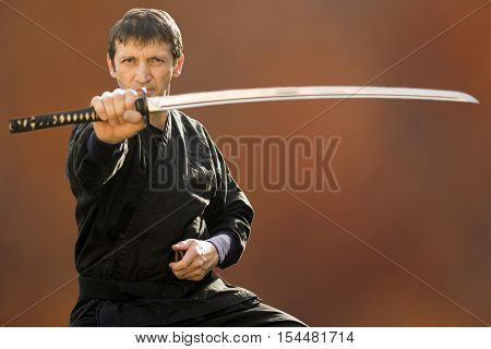 Sitting ninja in black kimono is holding his sword on brown background.
