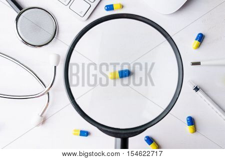 Close-up of magnifying glass and medicine with stethoscope keyboard and mouse. Top view of white wooden working table.