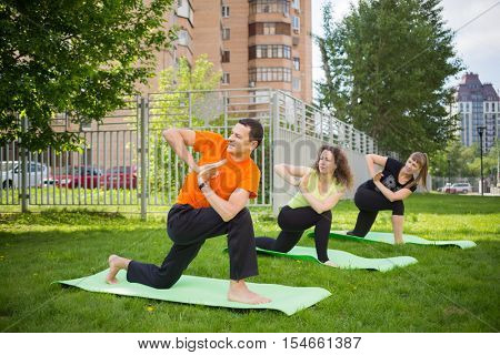 Man and two women practice yoga on grass near buildings at summer day