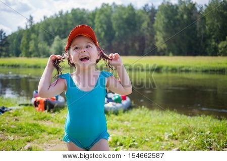 Little girl in swimsuit laughs near river with inflatable boats out of focus during camping trip at summer
