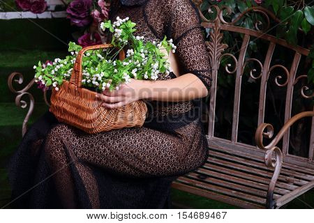 Woman in transparent dress holds basket with flowers on wrought iron bench in studio, noface