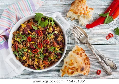 Chili con carne in a white casserole on wooden table with fresh vegetables and bread. Cooked with ground beef tomatoes peppers beans corn garlic onion cilantro. Top view.