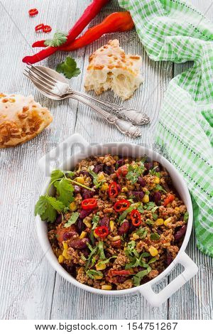 Chili con carne in a white casserole on wooden table with fresh vegetables and bread. Cooked with ground beef tomatoes peppers beans corn garlic onion cilantro.