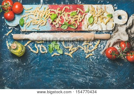 Ingredients for cooking Italian dinner. Fresh pasta casarecce, tomatoes, basil leaves, bottle of olive oil on colorful wooden board over dark blue background. Top view, copy space. Food frame concept