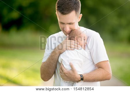 Portrait of young happy man holding his newborn cute babe dressed in white unisex clothing. Caucasian father embracing tenderly his adorable new born child and smiling. Outdoors shot