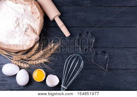 Ingredients for baking. Flour in paper bag wheat eggs and kitchen utensils - rolling pin and whisk on dark wooden background. Cooking cookies. Top view. Copy space