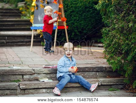 Group of two white Caucasian toddler children kids boy and girl outside in summer autumn park by drawing easel with markers apples books playing studying learning back to school