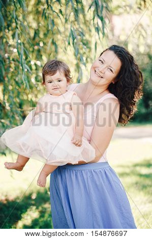 Lifestyle group portrait of smiling white Caucasian brunette mother holding hugging daughter in pink dress looking in camera on sunny spring summer day in park outside
