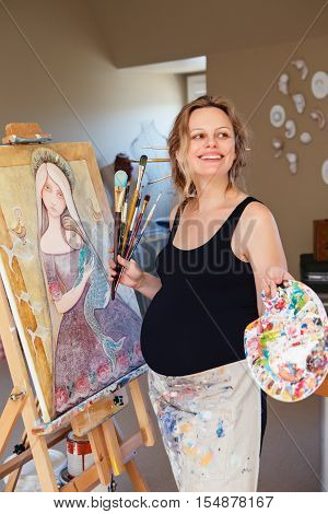 Portrait of smiling white Caucasian young pregnant woman drawing painting standing at easel in home studio art mental therapy healthy happy lifestyle concept