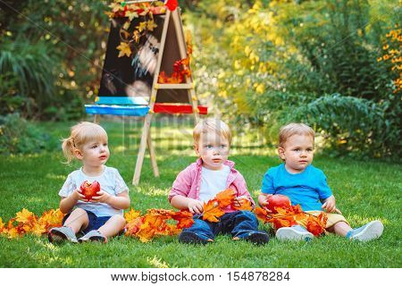Group of three white Caucasian toddler children kids boys and girl sitting outside in summer autumn park by drawing easel with fall leaves and apple back to school