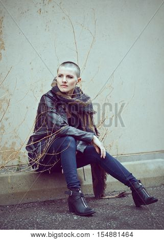 Portrait of beautiful Caucasian white young bald girl woman sitting on ground outside with shaved hair head in jacket scarf shaw jeans looking away toned with Instagram filters in blue green color