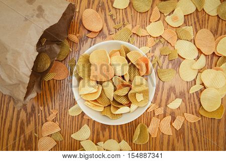 Closeup view from above of colorful veggie potato tomato spinach chips on a white plate and brown paper bag with greesy spots stains on a wooden table