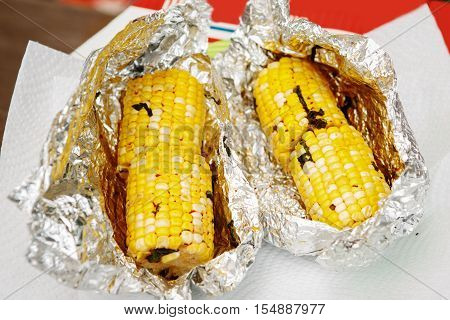 Two grilled sweet yellow corn cobs wrapped in foil paper on a table macro closeup