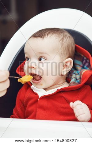 Close-up portrait of cute adorable Caucasian little baby boy with dark black eyes sitting in high chair in kitchen looking away eating meal puree mother feeds her son