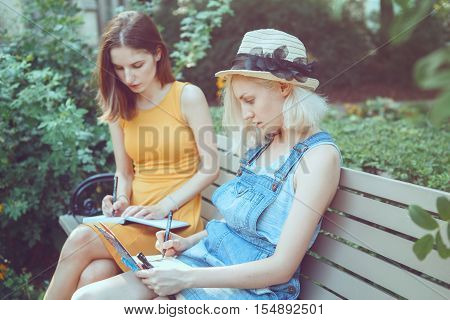 Portrait of two white Caucasian young girls hipster students friends outside in park on summer day sitting on bench together drawing sketching best friends forever toned with filters
