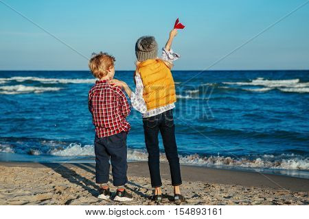 Group portrait of two white Caucasian children kids older sister and younger brother playing paper planes on ocean sea beach on sunset outdoors happy lifestyle childhood concept