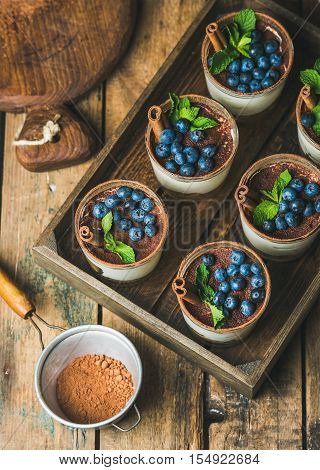 Homemade Tiramisu dessert in glasses with cinnamon, mint and fresh blueberries in wooden tray and sieve with cocoa powder over rustic wooden background, top view, selective focus, vertical composition