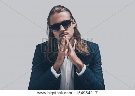 Young and full of confidence. Handsome young man in full suit keeping hands clasped and looking at camera while sitting against grey background