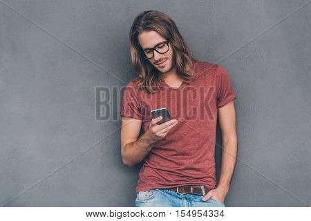 Typing quick message to friend. Handsome young man in casual wear holding smart phone and looking at it while standing against grey background