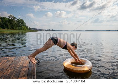 Side View Of One Young Girl Diving Into Water And Svim Ring From A Jetty At A Summer Lake.