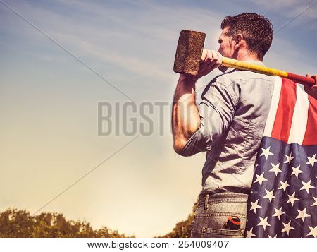 Attractive Guy With A Sledgehammer And A Us Flag On His Shoulders, Looking Into The Distance Against