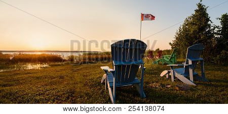 Muskoka Chairs On A Lawn Beside A Lake With A Canadian Flag In The Background At Sunset.