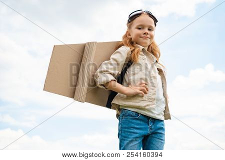Low Angle View Of Child With Paper Plane Wings On Back And Protective Eyeglasses On Head Against Blu