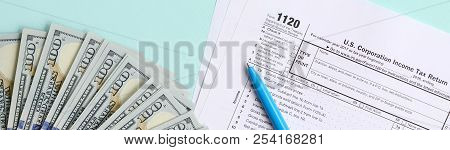 1120 Tax Form Lies Near Hundred Dollar Bills And Blue Pen On A Light Blue Background. Us Corporation