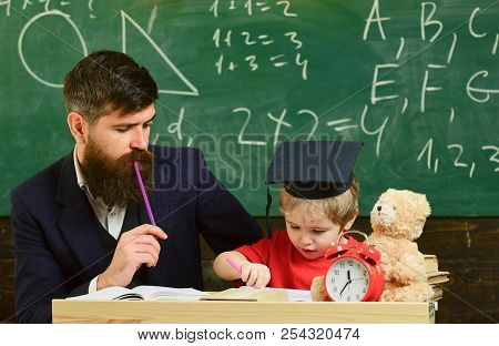 Father Checking Hometask, Helps To Boy, Son. Teacher In Formal Wear And Pupil In Mortarboard In Clas