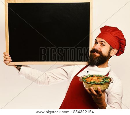 Catering And Open Day Concept. Cook With Tricky Smile In Burgundy Uniform Holds Baked Dish And Black