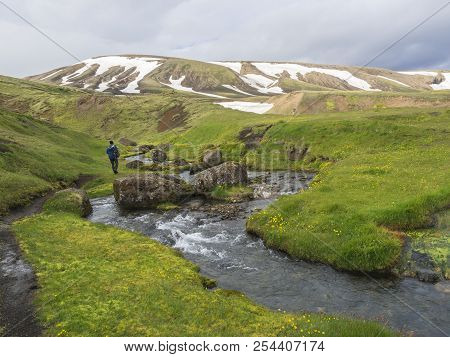 Lonely Man Hiker Walking In  Landscape With Wild Creek Stream, Green Grass Moss Meadow And Snow Cove