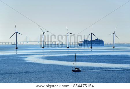 A Very Large Passenger Ship And A Small Sailboat Pass Offshore Wind Turbines Near The Oresund Bridge