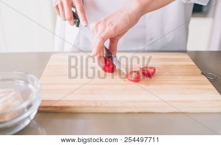 Close Up Of Woman's Hand Holding And Cutting Cerry Tomato. She Does It With Knife On Wood Board.