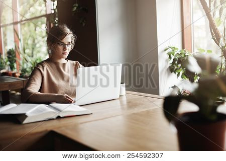 Attractve Female Entrepreneur Working With Laptop In Cafe, Surrounded With Cute Flowers, Wearing Gla