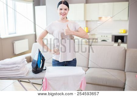 Positive Brunette Is Posing On Camera. She Holds Her Big Thumb Up And Smiling. There Is A N Iron And