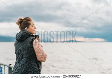 Outdoor Portrait Of Young Woman Resting By The Lake On A Fresh Cold Day, Wearing Warm Pullover And B
