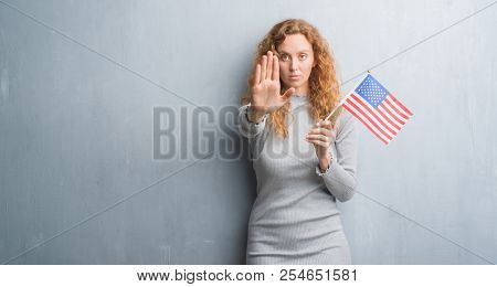 Young redhead woman over grey grunge wall holding flag of United States of America with open hand doing stop sign with serious and confident expression, defense gesture