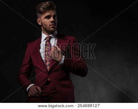 surprised young man in grena suit looks to side while fixing his collar and sitting on grey wallpaper background