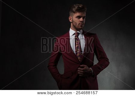 portrait of relaxed man buttoning his grena suit while looking to side and standing on grey wallpaper background