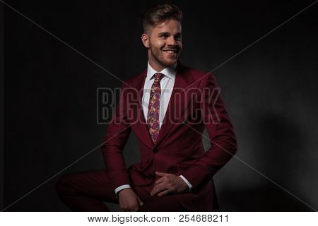 portrait of smiling elegant man in grena suit looking to side while sitting on wooden chair on grey background