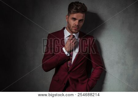 portrait of relaxed man in grena suit fixing his tie while standing on grey wallpaper background