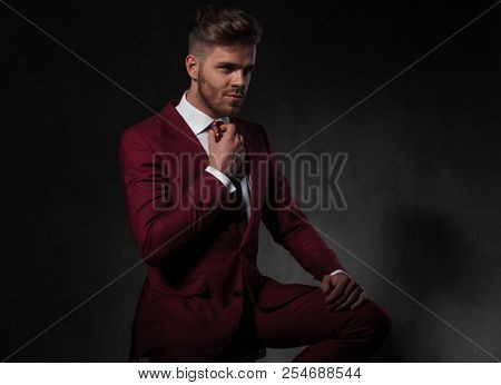 gentleman in suit fixing his red tie and looking to side while sitting on wooden stool on grey wallpaper background